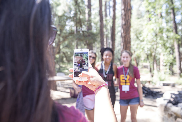 Two students posing for a photo with the photographer holding up a phone to take the photo in the foreground. In the background there are trees and greenery.