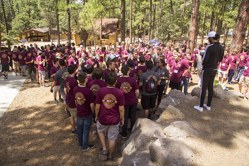 All the E2 campers (about 200) are shown outside in a large open area surrounded by forests participating in a grouping activity.