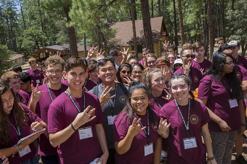A group of about 50 campers stand together making the ASU pitchfork gesture smiling for the camera outside in the sun