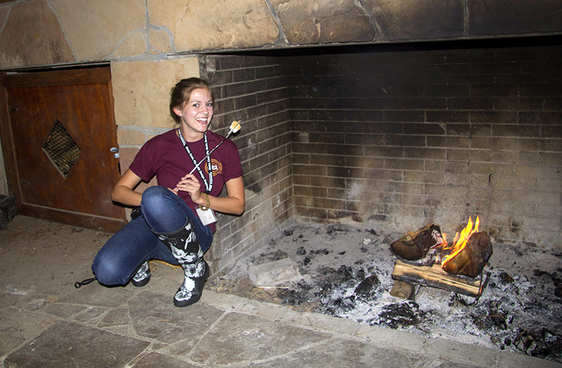 A young woman roasts a marshmallow in a large, rustic fireplace.
