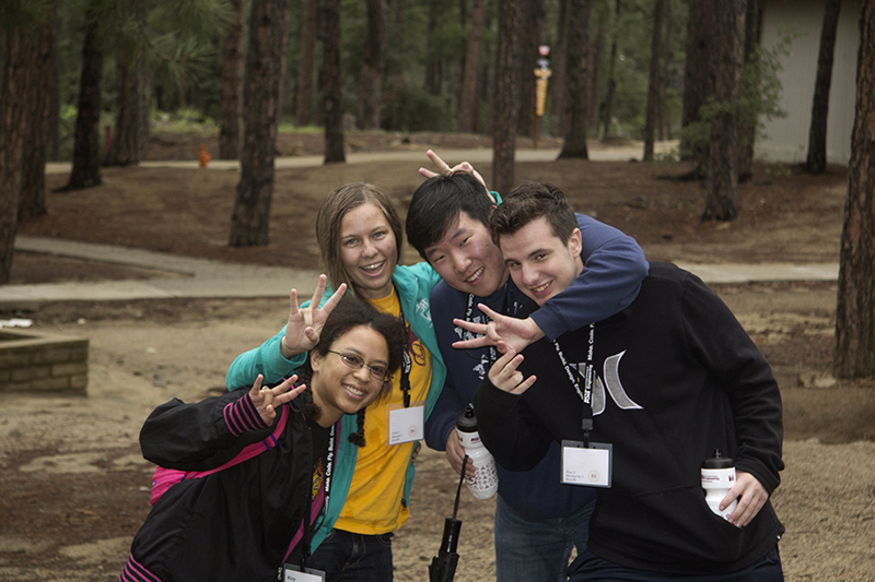 A group of 4 students, arms around one another, smile and lean in for a photo while making the ASU pitchfork gesture.